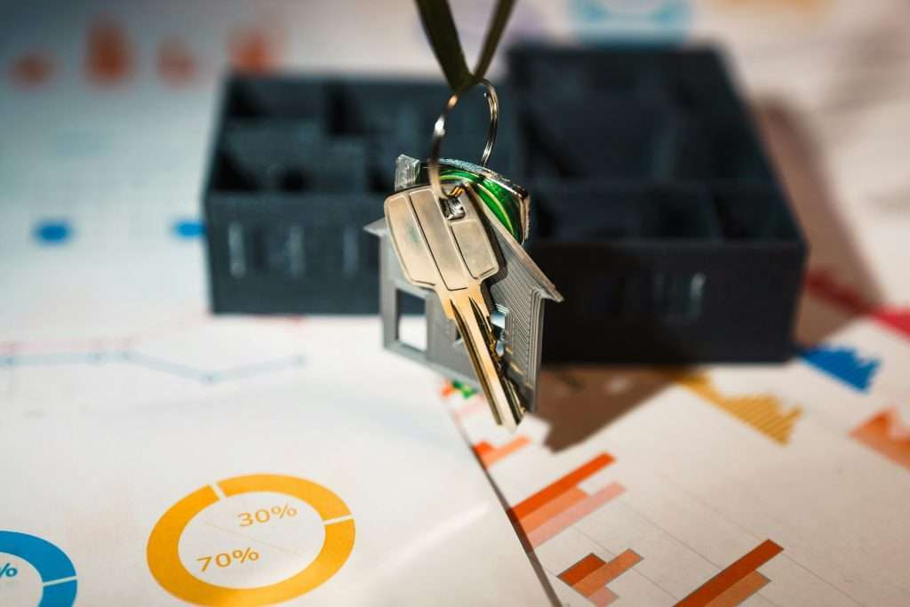 A close-up of a house-shaped keychain with keys, held above floor plans and colorful financial charts, symbolizing real estate investment or home buying.